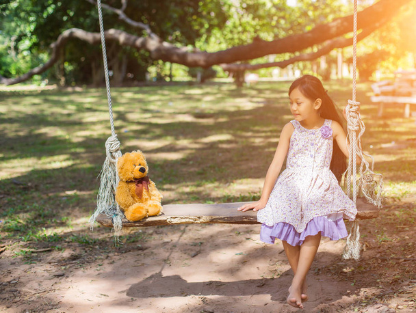 Roztomilá holčička sedí houpačky, medvídek sedí s ní, holčička ukazuje na strom. - Fotografie, Obrázek
