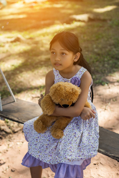 Küçük tatlı kız salıncakta sallanıyor, oyuncak ayı yanında oturuyor, küçük kız ağacı işaret ediyor.. - Fotoğraf, Görsel