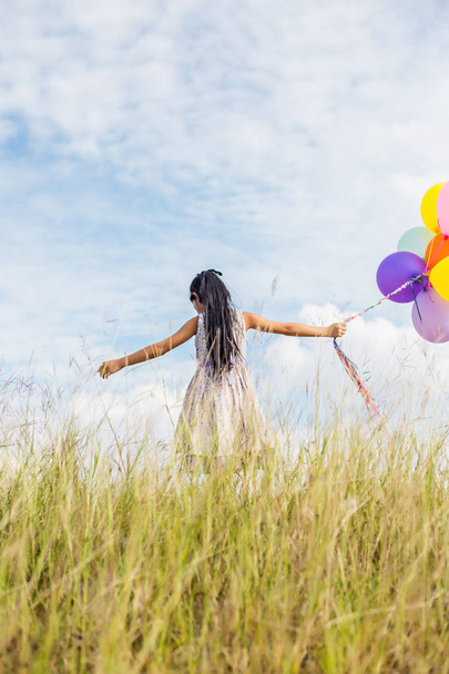 niedliches kleines Mädchen mit bunten Luftballons, die auf der Wiese vor blauem Himmel und Wolken laufen. - Foto, Bild