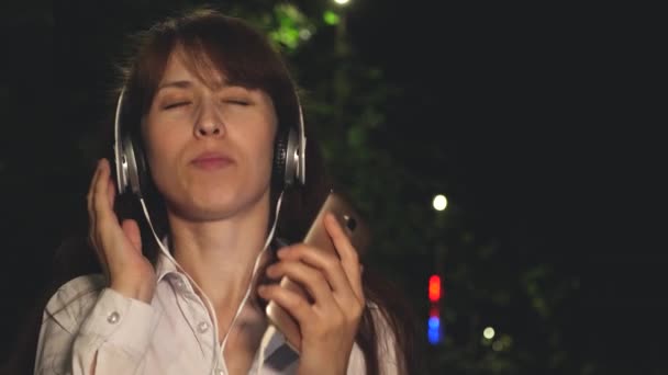 Girl in headphones and touch phone waving long hair singing and dancing smiling and kayfuya from music at night in park. Close-up - Materiaali, video