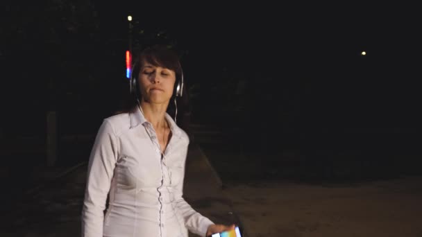 Młoda dziewczyna w słuchawkach z touch telefon tańce i śpiewa, uśmiechając się i coraz adrenaliny od muzyki w nocy w parku miejskim - Materiał filmowy, wideo