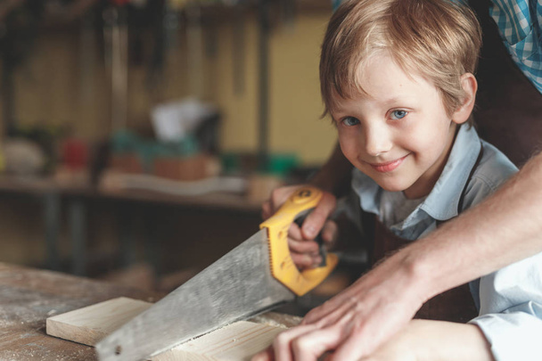 Pai e um pequeno filho a serrar uma tábua na carpintaria
 - Foto, Imagem