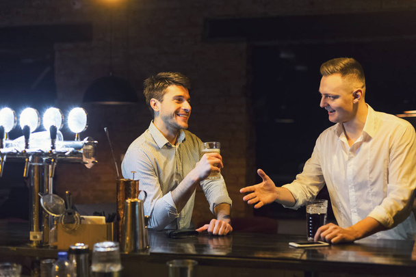 Dos jóvenes felices hablando y bebiendo cerveza en el bar
 - Foto, Imagen