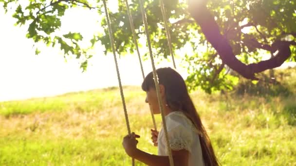 Beautiful girl with long hair swinging on swing in bright golden sunset and smiling. Slow motion - Footage, Video