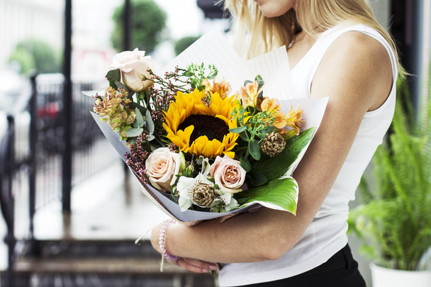 Young woman with blonde hair holding bouquet of flowers rose sunflower on the street - Photo, Image