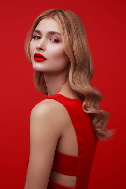 Studio shot van modieuze blonde vrouw in rode jurk. Hollywood kapsel en rode lippen. rode nagels. krullend modemodel met glamour make-up. jonge vrouw op rode achtergrond.  - Foto, afbeelding