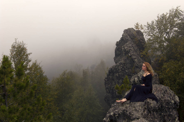 a vörös hajú lány mezítláb fekete ruha ül egy szikla fölött az erdő, a háttérben a közeledő fala köd (a rock és a csúcs a Mount Kolpaki a feltételes határ Európa és Ázsia középső Ural) - Fotó, kép