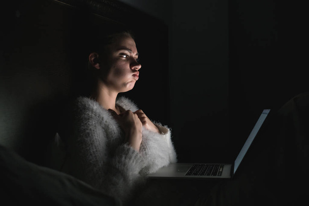 scared young girl in pajama sits in bed late at night, watching horror film on laptop - Zdjęcie, obraz
