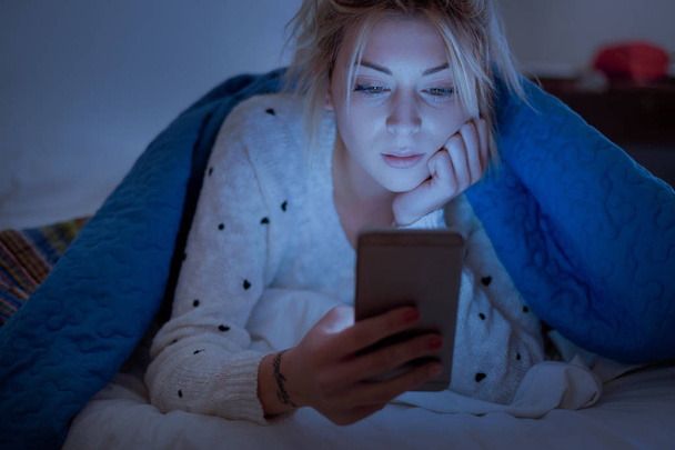 Disappointed sad woman holding mobile phone while laying on bed at night - Photo, Image