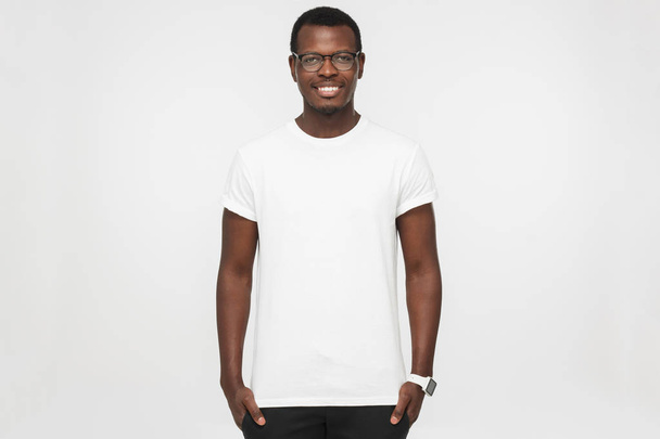 Handsome african american man wearing blank white t shirt, smart watches and trendy eyeglasses, isolated on grey background, smiling, standing in hands in pockets pose - Fotó, kép