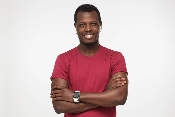 Young smiling african american man with crossed arms, wearing wireless headphones, red t shirt and smart watches with touch screen, isolated on gray background  - Φωτογραφία, εικόνα