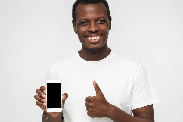 Closeup shot of young African American man isolated on white background wearing white t shirt, showing blank phone screen and thumb up willing to recommend product or service - Foto, Imagem