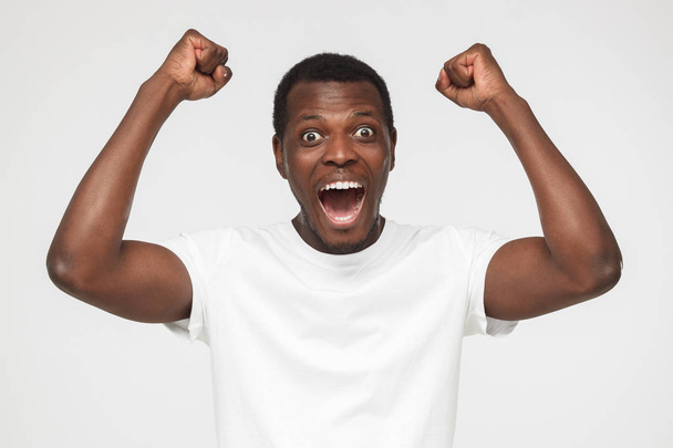 Young african american man in blank white t shirt, shouting while his team win, raised both fist in victory gesture, isolated on gray background - Foto, Bild