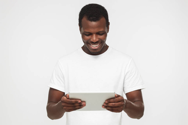Studio shot of young African American man pictured isolated on gray background wearing blank white t shirt looking at screen of tablet he is holding, smiling happily - Photo, image
