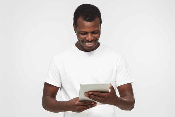 Portrait of young handsome african american man in blank white t shirt isolated on gray background looking at screen of tablet pc computer, reading interesting article  - Foto, immagini