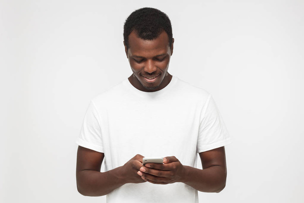 Young african american hipster man in blank white t shirt, isolated on gray background, texting or browsing with happy relaxed smile - Foto, immagini