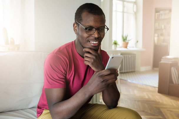 Indoor photo of handsome African man dressed in casual clothes spending time in living room holding mobile phone looking at screen through glasses with calm smile receiving good news from web - Zdjęcie, obraz