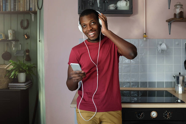 Horizontal shot of African American man standing in his kitchen in casual clothes with big headphones on, listening to music from his phone smiling happily and enjoying his free time at home - Foto, afbeelding