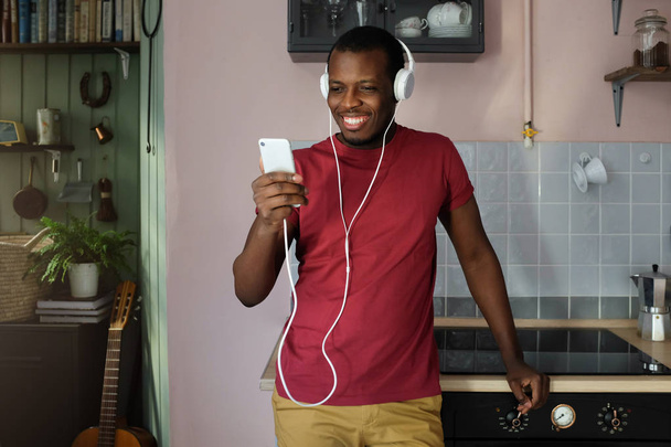 Indoor picture of young African American man staying at home, listening to his favorite media files on his phone while standing in kitchen, using trendy headphones, looking satisfied - Foto, afbeelding