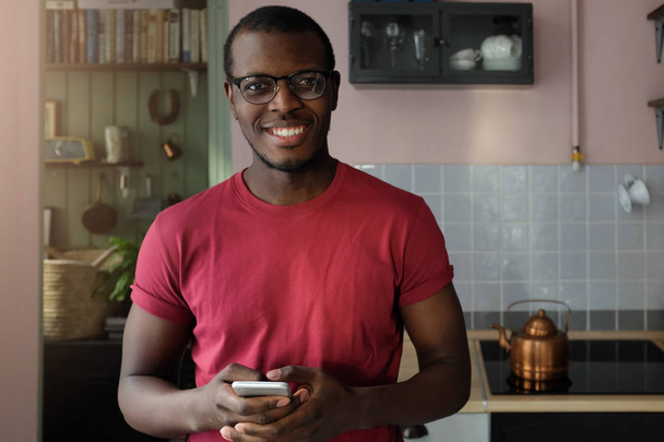 Horizontal closeup shot of young African American man spending afternoon in his kitchen at home looking at camera through glasses with smile, using cellphone to communicate or browse websites - Фото, зображення