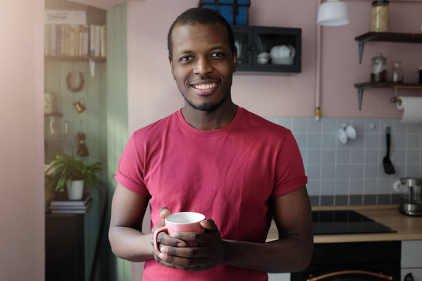 Indoor closeup photo of young African man standing in colored T-shirt in his kitchen smiling friendly, looking at camera, holding coffee cup in hands during his breakfast, enjoying free morning - Foto, imagen