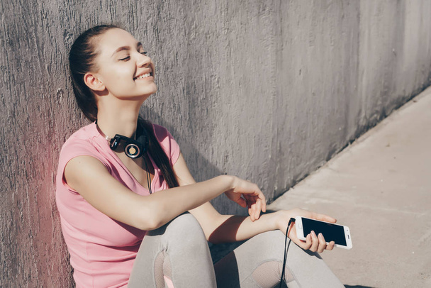 happy girl in pink t-shirt is sitting on the ground, resting after jogging and smiling - 写真・画像