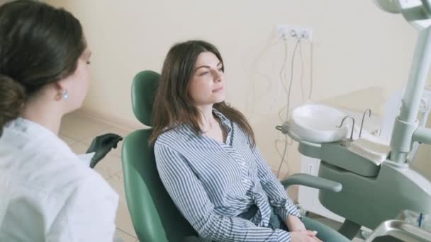 A beautiful smiling brunette with long hair in stylish clothes came to treat her teeth in the dentists office. Slowly shooting steadikam the doctor puts an apron on the patients chest, prepares her - Кадри, відео