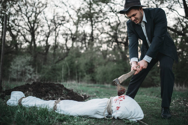 Маньяк с топором против тела жертвы, завернутого в холст, концепция серийного убийцы, ужас преступления
 - Фото, изображение