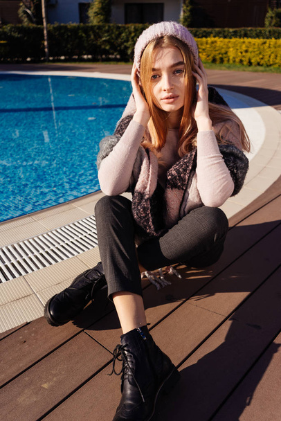stylish young blonde girl in hat and warm clothes is resting in the sun by the blue pool, looking at the camera - Fotoğraf, Görsel