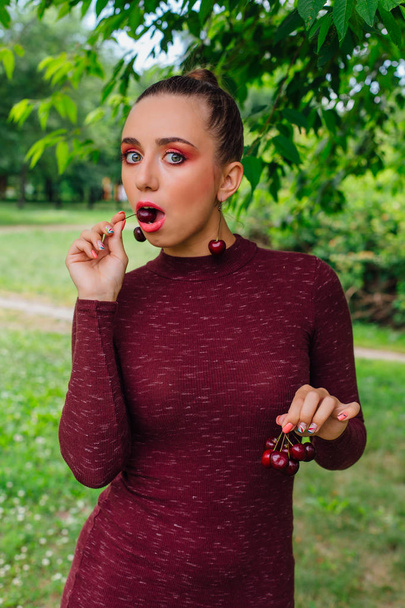 Beautiful young woman with long braid and cherry earrings holding cherry in hands - Foto, imagen