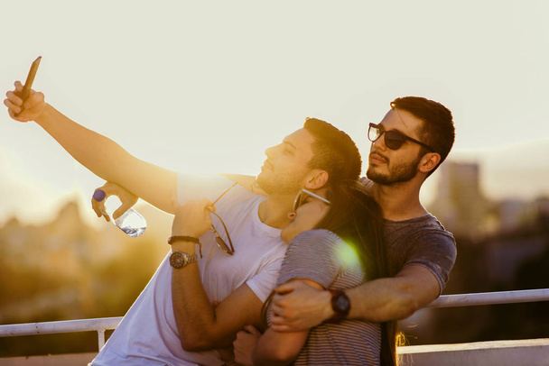 Dois amigos tirando selfies ao ar livre no terraço ao pôr do sol com uma luz quente no fundo
 - Foto, Imagem