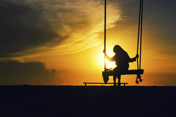 Silhouet van eenzaam meisje met hijab op een schommel met magische zonsopgang achtergrond. - Foto, afbeelding