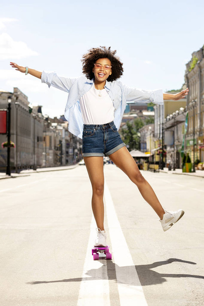 Cheerful curly girl posing while standing on mini skateboard - Zdjęcie, obraz