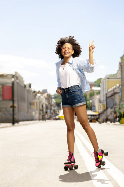 Happy curly girl showing V sign while roller-skating - Photo, Image