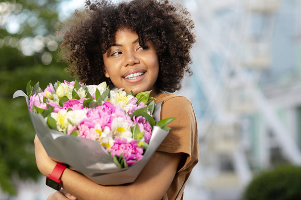 Best gift. Charming curly woman smiling widely and looking into the distance while holding a bouquet of flowers - Foto, Imagen