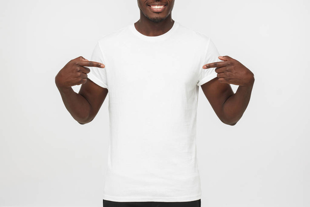 Smiling nice african american man pointing at blank white t-shirt with both index fingers, copy space for your advertising, isolated on grey background - Foto, imagen