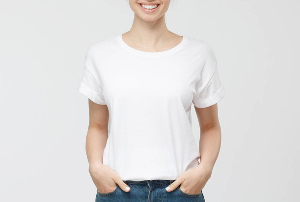 Young european woman standing with hands in pockets, wearing blank white tshirt with copy space for your logo or text, isolated on grey background - Photo, Image