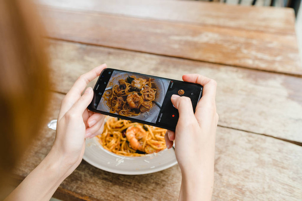 Vrouwelijke blogger fotograferen lunch in restaurant met haar telefoon. Een jonge vrouw nemen foto van spaghetti eten op smartphone, maaltijd fotograferen met mobiele camera. - Foto, afbeelding