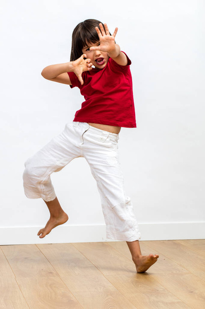 excited sporty young child with dynamic gesture fighting, showing energetic grace and power with dancing legs and hands for kid's martial art over wooden floor, white background - Фото, изображение