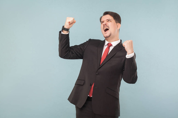 Happy successful businessman with mustache wearing black suit and red tie with raised hands rejoicing victory on light blue background   - Foto, afbeelding