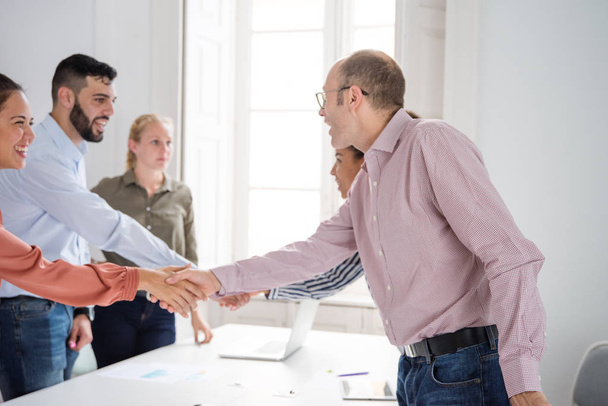 Business team in an office standing around a table shaking hands and greeting each other while they are smiling - Fotoğraf, Görsel