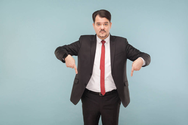 businessman with mustache wearing black suit and red tie looking at camera and pointing fingers down on light blue background - 写真・画像