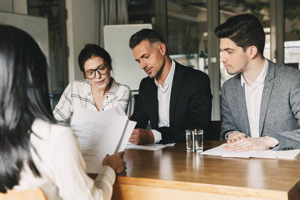 Business, career and recruitment concept - team of employers in formal wear sitting at table in office and interviewing woman for job in big corporation - Photo, Image