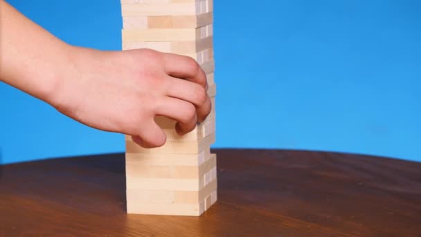 hands of Young man plays jenga on blue background, close-up. A man builds a tower of blocks while playing jenga - Felvétel, videó