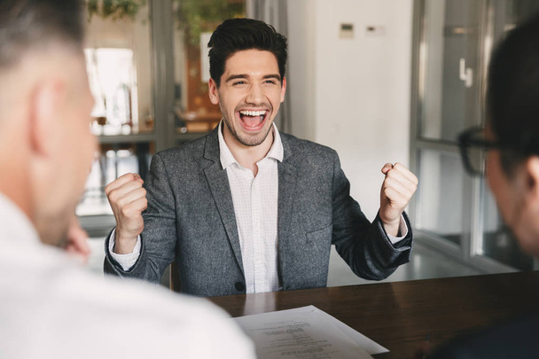 Business, career and placement concept - successful caucasian man 30s rejoicing and clenching fists during job interview in office with employees of big company - Photo, image