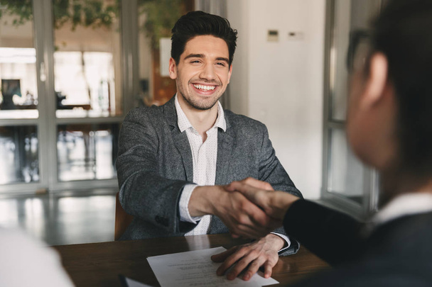 Business, career and placement concept - happy caucasian man 30s rejoicing and shaking hands with employee when was recruited during interview in office - Photo, image