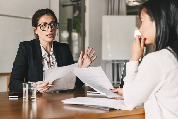 Photo of strict caucasian woman holding resume and negotiating with female candidate during corporate meeting or job interview - business, career and placement concept - Photo, image