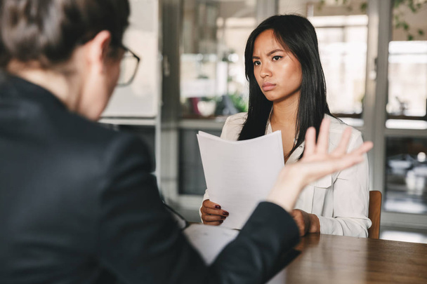 Image of serious asian woman looking and talking to businesswoman while sitting at table in office during job interview - business, career and recruitment concept - Foto, Imagem
