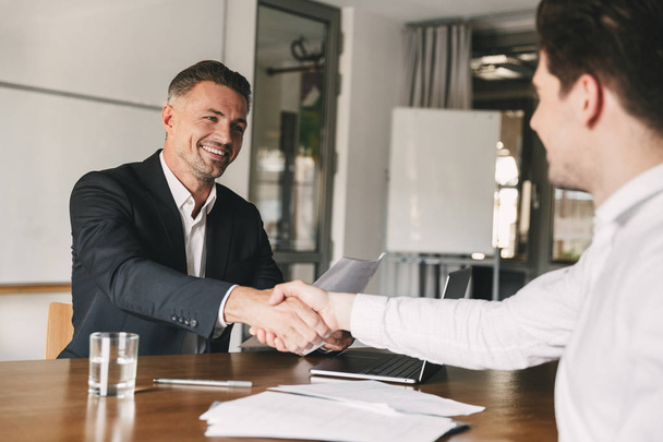 Business, career and placement concept - joyful handsome businessman 30s smiling and shaking hands with male candidate who was recruited during interview in office - Photo, image