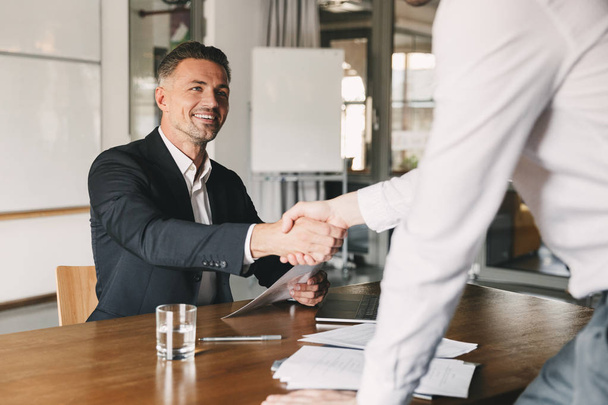 Business, career and placement concept - satisfied director man 30s smiling and shaking hands with male candidate who was recruited during interview in office - Photo, Image